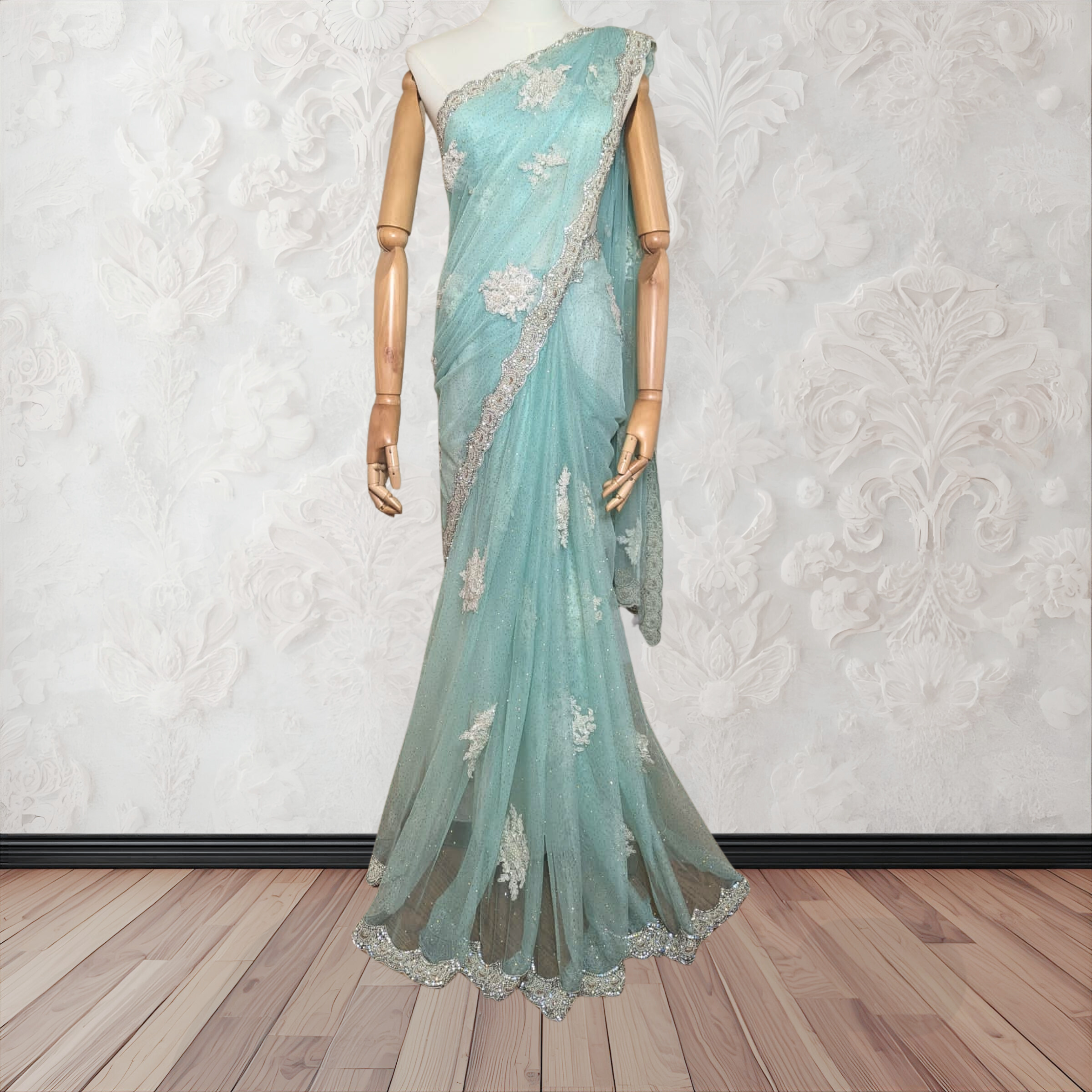 Teal saree with silver embroidery on a mannequin against a white textured wall.